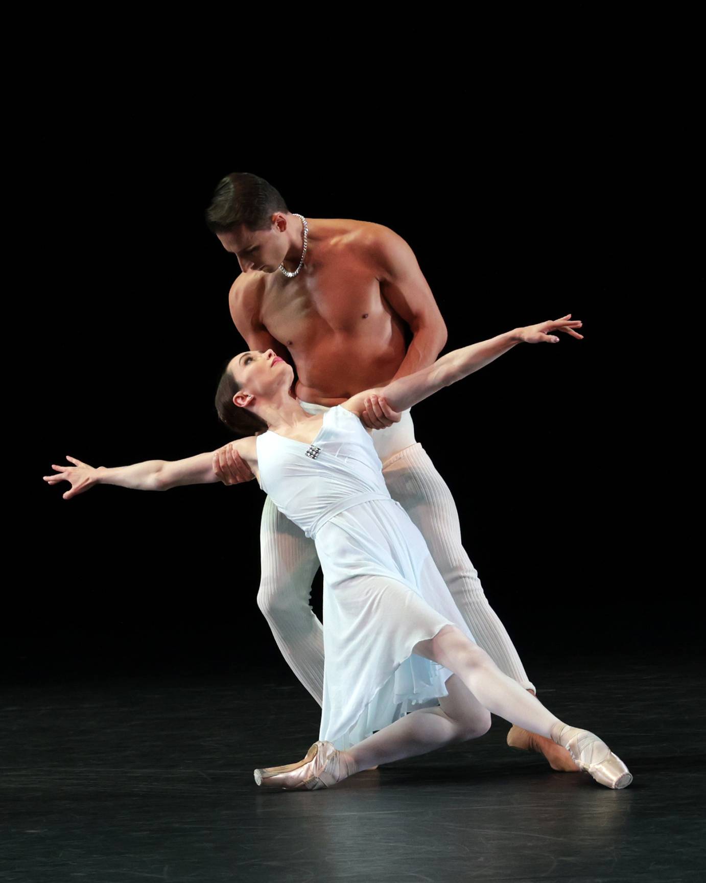 A barechested man holds a woman under her arms as he lifts her from the ground. They gaze into each other's eyes